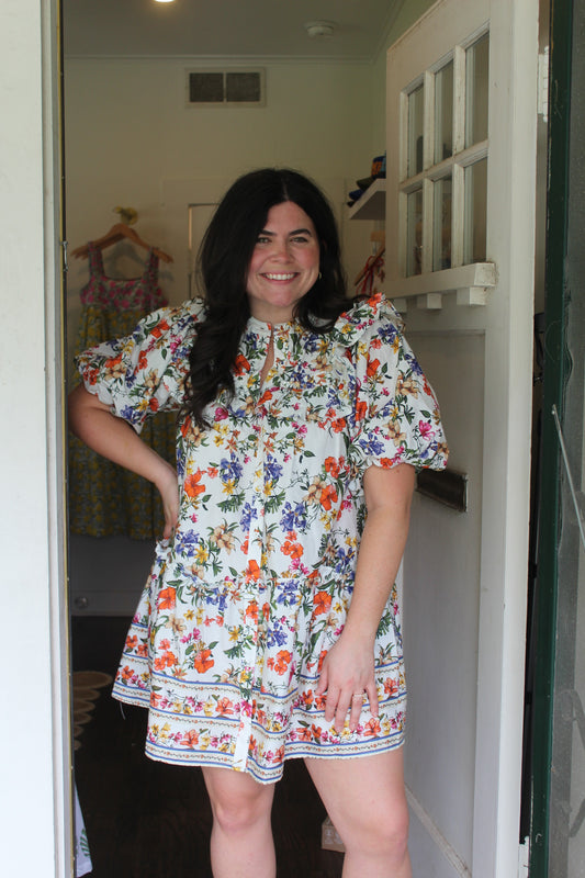 Model wearing the Elyse Ruffle Dress, a white floral mini with ruffle sleeves, in a doorway