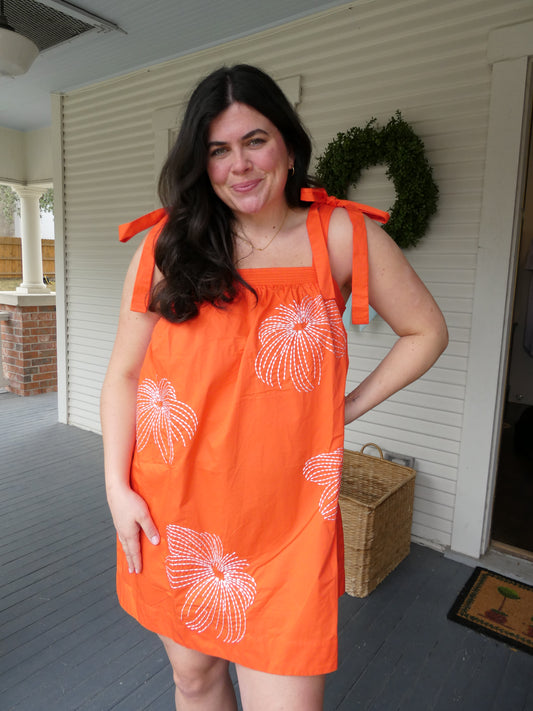 Model wearing the Viva Dress, an orange dress with white floral stitching and bow straps