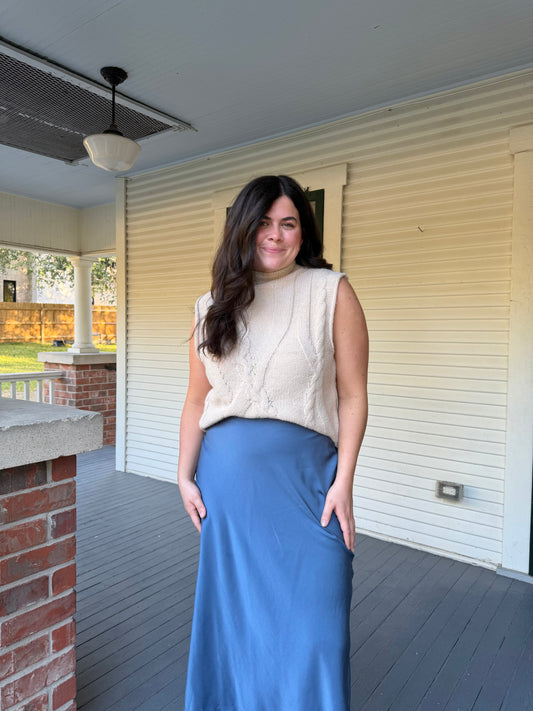 Model wearing the Sleeveless Sweater, an ivory cable knit, with a blue satin skirt