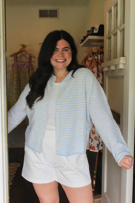 Model wearing the Light Blue Stripe Piper Cardigan open over white shorts in a doorway