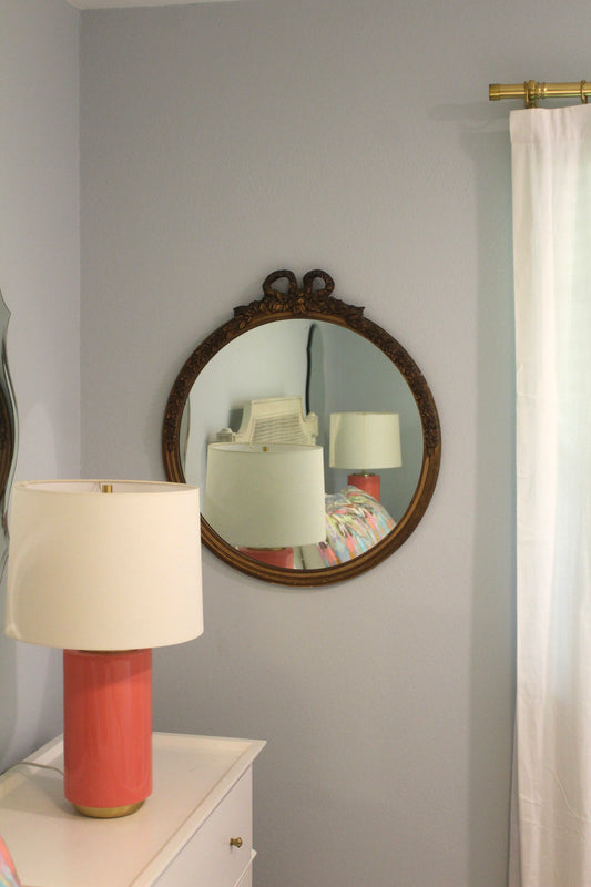 Vintage bow mirror hanging on a wall above a white dresser with a coral lamp